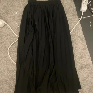 Svart långkjol med resårmidja - En svart långkjol med mjukt fall och resår i midjan. Kjolen har en klassisk och enkel design som gör den lätt att matcha med olika toppar. Perfekt för dig som gillar en stilren och tidlös look.