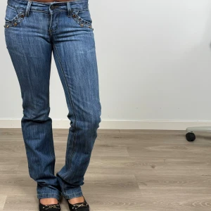 Blå jeans med broderade bakfickor - Snygga blå jeans från Big Bell  med broderade detaljer och paljetter på bakfickorna. Jeansen har normal passform, raka ben och dekorativa nitar framtil med coola detaljer som sticker ut. Storleken är W 27 L 32