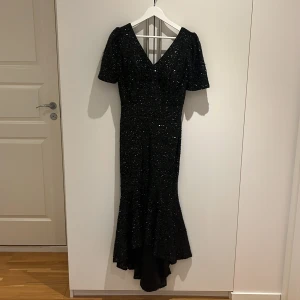 Svart glittrig långklänning med v-ringning - Supersnygg svart långklänning med glittriga paljetter över hela klänningen. Djup v-ringning fram och bak, korta ärmar och figurnära passform som avslutas med en volang nertill. Perfekt för dig som vill sticka ut på bal eller fest!