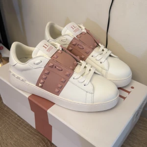 Valentino Garavani Rockstud sneakers - Snygga vita Valentino Garavani Rockstud sneakers med breda ljusrosa band och ikoniska nitar. Skorna har rund tå, platt sula och är gjorda i lyxigt skinn. Perfekta för dig som vill sticka ut med en exklusiv touch och coola detaljer.