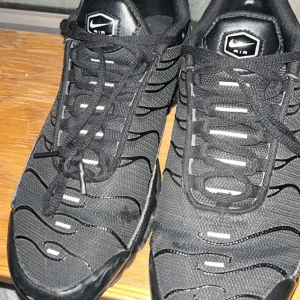 Nike Air Max Plus Tn Svarta sneakers - Nike Air Max Plus Tn sneakers i helsvart med coola vågformade detaljer och synlig Air-dämpning i sulan. Skorna har snörning, mesh och syntetmaterial på ovandelen samt Tn Air-logga bak. Perfekt för dig som gillar streetwear och sportig stil.
