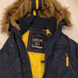 Mörkblå parkas från Alpha Industries - Parkas från Alpha Industries i mörkblått med gul insida och detaljer. Jackan har fuskpäls på huvan, flera fickor med dragkedja och knappar samt patch på ärmen. Perfekt för kalla dagar med sin långa modell och fodrade insida.