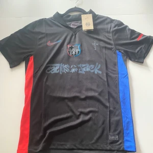 FC Barcelona x Cactus Jack Lamine Yamal - Snygg svart FC Barcelona fotbollströja med Cactus Jack-grafik på bröstet och Lamine Yamal 10 på ryggen. Tröjan har röda och blå sidopaneler, klubbmärke på bröstet och UNHCR-tryck bak. Tillverkad i lätt och ventilerande material.