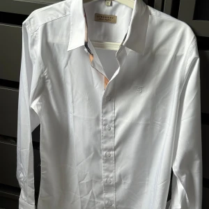 Vit Burberry skjorta  - Klassisk vit skjorta från Burberry i storlek M, med diskret broderad logga på bröstet. Skjortan har långa ärmar och knappar framtill. Kragen och ärmsluten har snygga rutiga detaljer i beige och rött som ger en lyxig känsla. Perfekt för dig som gillar stilrena plagg med exklusiva inslag.