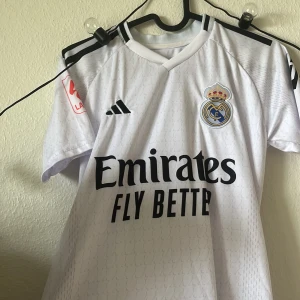 Real Madrid Bellingham matchtröja 164 - Säljer en vit Real Madrid fotbollströja från Adidas med Bellingham och nummer 5 på ryggen. Tröjan har korta ärmar, svarta detaljer och klubbmärke på bröstet. Materialet är lätt och andas, perfekt för fotbollsträning eller match.