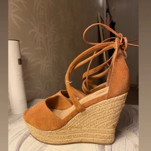 UGGS, klackar 🤎 - Snygga sandaler från UGG i brun mocka med höga flätade kilklackar och knytband runt vristen. Skorna har öppen tå och plattformsula, vilket ger en somrig och trendig look. Perfekta till både jeans och klänning för en avslappnad stil. Skorna är i gott skick förutom en liten fläck på ena skons framsida som säkert går att tvätta av! Storlek 38. 