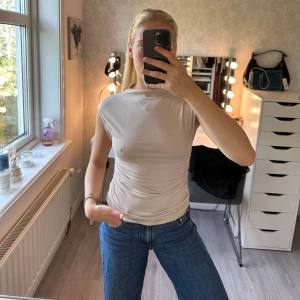 Beige ärmlös topp med rynkade sidor - Snygg beige topp med ärmlös design och hög hals. Toppen har rynkade detaljer längs sidorna som ger en cool, figursydd och smickrande passform. Tillverkad i ett mjukt och stretchigt material som känns skönt mot huden. Aldrig använd🤎