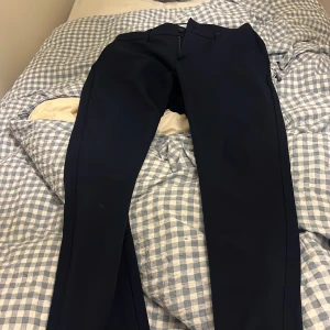 Midnattsblå kostymbyxor från Woodbird - Snygga midnattsblå kostymbyxor från Woodbird i modellen Steffen Twill Pant. Byxorna har raka ben, bälteshällor, knäppning med dragkedja och knapp samt två bakfickor med knapp. Perfekta för en clean och stilren look.