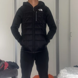 Svart puffer väst från The North Face - Svart pufferjacka från The North Face med huva och dragkedja framtill. Jackan har quiltad front och rygg, samt släta ärmar. Bröstficka med dragkedja och logga på bröstet och ryggen. Perfekt för kyliga dagar och riktigt snygg streetstil.