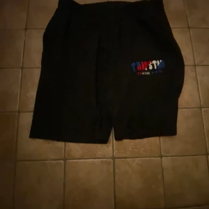 Svarta Trapstat shorts med brodyr - Svarta shorts från Tripstar med färgglad broderad logga på ena benet. Klassisk passform med elastisk midja, perfekta för chill dagar eller streetwear. Materialet känns mjukt och bekvämt, och designen är enkel men ändå snygg.