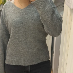 Grå stickad tröja med v-ringning - Mysig grå stickad tröja med v-ringad hals och långa ärmar. Tröjan har en enkel och stilren design med ribbade muddar vid ärmslut och nederkant. Perfekt för lager-på-lager och chill dagar.