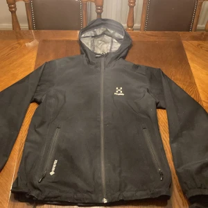 Svart vindjacka från Haglöfs S - Svart vindjacka från Haglöfs i storlek S. Jackan har huva, dragkedja framtill, två sidofickor med dragkedja och meshfoder på insidan. Tillverkad i slitstark polyester med GORE-TEX-teknologi för extra väderskydd. Perfekt för uteliv och äventyr.