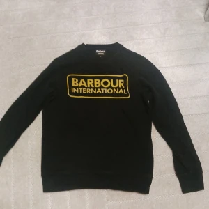 Svart Barbour International sweatshirt - Svart sweatshirt från Barbour International med stor gul logga framtill. Klassisk rund halsringning och långa ärmar. Tröjan är i mjuk bomullsmix och har en avslappnad passform som funkar till det mesta.