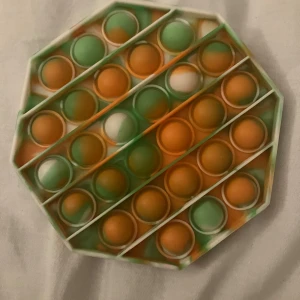 Pop It Fidget Toy - Färgglad Pop It fidget-leksak i silikon med orange, gröna och vita nyanser. Åttakantig form, perfekt för stresslindring och sensorisk stimulans. Inga synliga defekter eller slitage, i mycket gott skick.