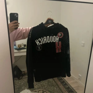 Svart hoodie från Hoodrich med tryck - Svart hoodie från Hoodrich i storlek S med stora vita och röda tryck framtill samt broderade detaljer. Klassisk huva och långa ärmar, perfekt för en streetwear-look. Mjuk insida och ribbade muddar för extra komfort.