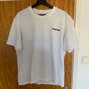 Vit Patagonia t-shirt med tryck - Vit t-shirt från Patagonia i bomull med klassiskt logotryck på bröstet och stort färgglatt Patagonia-tryck på ryggen. Regular fit och rund hals. Perfekt för dig som gillar stilrena och ikoniska plagg. Har du fler frågor får du gärna höra av dig. Priset kan diskuteras vid snabb affär.