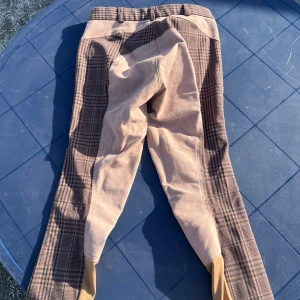 Rutiga ridbyxor från JR Collection - Snygga ridbyxor i brunt och beige från JR Collection med rutigt mönster och kontrastfärgade detaljer vid fickor och benslut. Perfekta för dig som gillar stil på ridbanan.