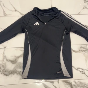 Svart Adidas träningströja med dragkedja - Svart långärmad träningströja från Adidas med vita detaljer och klassiska ränder på ärmarna. Slim fit-modell med halv dragkedja framtill och hög krage. Tillverkad i lätt polyester som andas, perfekt för sport eller gym.  Tyckte att den var för tajt men glömde skicka tillbaka