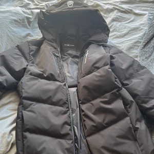 Svart Sail Racing dunjacka med huva - Svart dunjacka från Sail Racing med stor huva och Sail Antarctica Expedition-märke på ärmen. Jackan är vadderad med certifierat dun (500 FP), har dragkedjor framtill och på bröstfickan samt är tillverkad i återvunnet material. Perfekt för kalla vinterdagar.