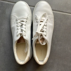 Vita sneakers från Gant i skinn - Snygga vita sneakers från Gant i skinn med klassisk design. Skorna har rund tå, platt sula och snörning. Beige detalj på hälen och diskret Gant-logga på sidan och baksidan. Perfekta för en clean och stilren look.