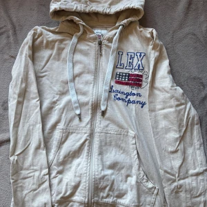 Beige hoodie från Lexington Company - Snygg beige hoodie från Lexington Company med dragkedja, huva och broderad amerikansk flagga samt text på bröstet. Tröjan har känguruficka och snörning i huvan. Perfekt för en avslappnad och sportig stil.