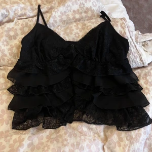 Svart volangig spets-linnetopp - Säljer en svart linne-topp med tunna axelband och flera lager volanger i spets och tyg. Toppen har en feminin och cool look med detaljerad spets och volanger som ger extra attityd. Perfekt för dig som gillar att sticka ut med din stil.