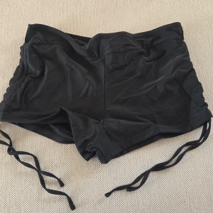 Svarta shorts med snörning i sidan - Svarta shorts i stretchigt material med snygg snörning i sidorna. De är korta och har en tight passform, perfekta för träning eller dans. Materialet är mjukt och följsamt, tillverkat av nylon och spandex för maximal rörelsefrihet. Använda en gång.