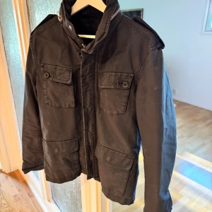 Svart parkajacka med dragkedja S - Svart parkajacka med fyra stora fickor framtill, axelklaffar och hög krage med dragkedja och knappdetaljer. Jackan har en rak passform och är tillverkad i bomullsmaterial. Perfekt för dig som gillar en clean och funktionell stil. Passar nu perfekt inför hösten och om du har några funderingar är det bara att höra av sig🫡