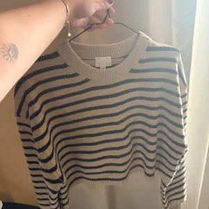 Randig stickad tröja i beige och svart - Säljer en snygg stickad tröja med beige bas och svarta ränder. Tröjan har en croppad passform, rund halsringning och långa ärmar. Perfekt för lager-på-lager och passar till både jeans och kjol.