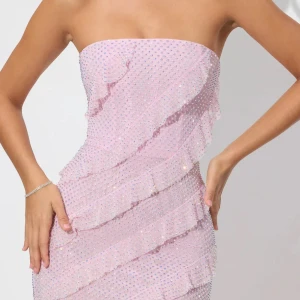 Ljusrosa glittrig långklänning - Säljer en ljusrosa axelbandslös långklänning med glittriga paljetter och volangdetaljer som går diagonalt över hela klänningen. Klänningen har en figurnära passform och är perfekt för dig som vill sticka ut med en glamorös look. SÄLJS INTE LÄNGRE 