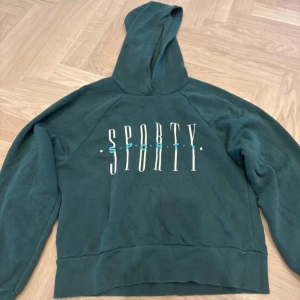 Grön hoodie SPORTY från Gina Tricot XS - Mörkgrön hoodie från Gina Tricot i storlek XS med trycket 'SPORTY' i vitt och blått framtill. Tröjan har huva och är tillverkad i mjuk bomullsblandning, perfekt för en chill och sportig look.