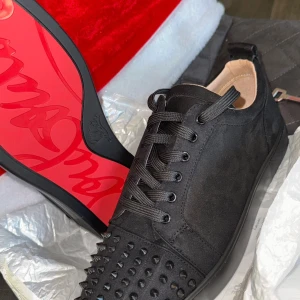 Christian Louboutin svarta sneakers med nitar - Säljer ett par svarta sneakers från Christian Louboutin med ikonisk röd sula och coola svarta nitar framtill. Skorna har snörning och är tillverkade i mocka för en lyxig känsla. Perfekta för dig som vill sticka ut med en edgy och exklusiv stil.