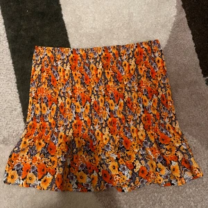 Blommig volangkjol i orange och gult - Kort kjol med blommigt mönster i orange, gult och inslag av svart och lila. Kjolen har volang nertill och en smockad midja som ger en bekväm passform. Perfekt för dig som vill sticka ut med färg och mönster.