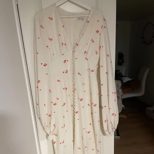 Vit blommig långklänning från Adoore - Superfin långklänning från Adoore i vit ton med små röda blommor. Klänningen har v-ringning, långa puffärmar och knappar framtill. Materialet är lätt strukturerat och känns luftigt. Perfekt för sommarens alla tillfällen!