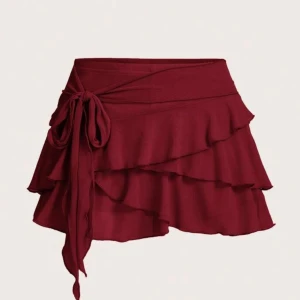 Vinröd omlottkjol med volanger - Supersnygg vinröd kortkjol med omlottmodell och volanger. Kjolen har en knytning i midjan som ger en extra detalj och en feminin look. Perfekt för dig som vill sticka ut med en trendig och lekfull stil. Inbyggda mjuka shorts 