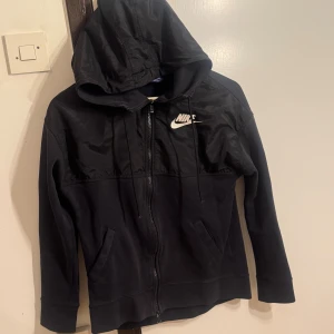 Svart Nike hoodie med dragkedja - Svart hoodie från Nike med vit logga på bröstet. Tröjan har huva med snörning, dragkedja framtill och två fickor. Överdelen är i glansigt material medan resten är i mjuk bomull. Perfekt för en sportig och avslappnad stil.