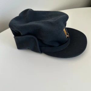 Mörkblå militärkeps med emblem - Säljer en mörkblå militärkeps med klassisk form och styv skärm. Framtill sitter ett guld- och blått emblem med tre kronor. Kepsen är tillverkad i ett kraftigare tyg, troligen ull, och har en snygg, tidlös look.