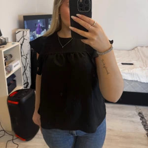 Svart blus med volangärmar - Säljer en svart blus med fyrkantig halsringning och volangdetaljer på axlarna. Blusen är ärmlös och har en lös passform som ger en avslappnad men ändå stilren look. Perfekt att matcha med jeans eller kjol.