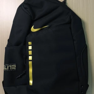 Svart Nike Elite ryggsäck med guldtryck - Snygg svart Nike Elite ryggsäck med guldiga detaljer och logga. Ryggsäcken har flera fack, vadderade axelremmar med guldtext och transparenta paneler. Perfekt för dig som vill ha en sportig och stilren väska med mycket plats. Ryggsäcken är helt ny utan etikett. Pris kan diskuteras. 