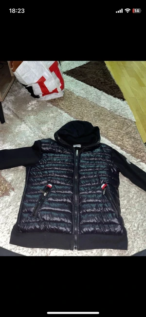 Svart pufferjacka från Moncler S - Svart pufferjacka från Moncler i storlek S. Jackan har quiltad framsida i glansigt material, mjuka svarta ärmar och huva. Två fickor med dragkedja och Moncler-logga på vänster ärm. Snygg och sportig look med detaljer i rött och vitt vid fickorna.