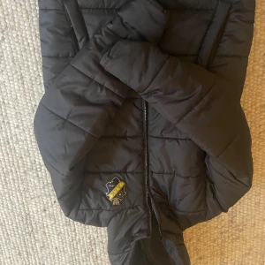 Svart AIK pufferjacka XS - Svart pufferjacka från AIK med klubbens emblem i gult och svart på bröstet. Jackan har huva, dragkedja framtill och quiltad design. Perfekt för dig som vill visa ditt AIK-supporterhjärta och hålla dig varm under kalla dagar. En liten reva bak på axeln. Se bild. Finns på Södermalm eller Skytteholm. 