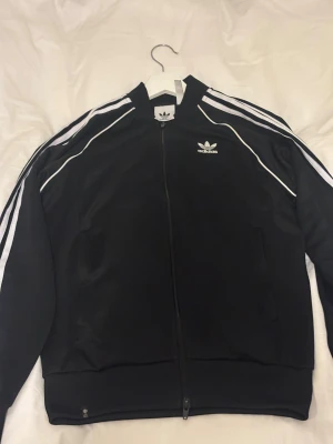 Svart Adidas bomberjacka med vita ränder - Klassisk svart Adidas bomberjacka med vita ränder längs ärmarna och broderad logga på bröstet. Jackan har dragkedja framtill, två fickor och ribbade muddar. Tillverkad i polyester och har en sportig, normal passform.