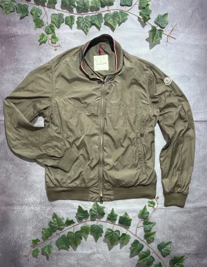 Moncler Albert Windbreaker i Mörkgrön. Storlek 5 (L/M) [ÄKTA] - 🎨 Färg: Mörkgrön / Olivgrön 📏 Storlek: Moncler 5 Vilket Motsvarar L/M ♻️ Skick: 10/10 – minimalt slitage, inga synliga skador eller fläckar 🧵 Material: Lättviktsnylon med ribbade muddar ✨ Detaljer: • Klassisk bombermodell • Hög krage med röd/vit kantband • Två framfickor med dragkedjor • Ribbstickade muddar vid midja och ärmslut • En knapp vid kragen för extra stängning • Moncler-patch på vänster överarm • Tvåvägsdragkedja ‼️ Skickas dagen efter köpet genomförs! ‼️