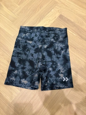 Mönstrade träningsshorts från 157 - Snygga träningsshorts från 157 med mörkblå och grå batikmönster. Modellen är högmidjad och har snörning i midjan för extra passform. Materialet är snabbtorkande polyester, perfekt för intensiva träningspass. Logga på benet och insidan.