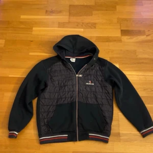 Svart Moncler med huva, öppen till byten - Svart bomberjacka från Moncler med quiltad front, huva och dragkedja. Jackan har ribbade muddar med röd, vit och blå rand, samt Moncler-logga på ärmen. Perfekt för dig som vill ha en stilren och sportig look. (det är en del av en dress, byxor finns också. skicka PM för dom)