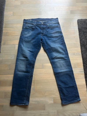 Blå jeans från G-Star RAW straight fit - Snygga blå jeans från G-Star RAW med klassisk femficksdesign och bruna detaljer på bakfickorna. Jeansen har en rak passform och mörk tvätt med lätt slitning framtill. G-Star RAW-logga på läderpatch bak i midjan. Perfekt för dig som gillar stilrena jeans. Jeansen är använda max 10 gånger
