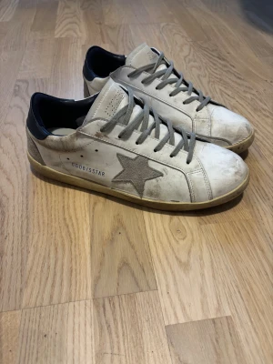 GGDB Superstar vita sneakers med stjärna - Säljer ett par GGDB Superstar sneakers i vitt skinn med grå mockadetaljer och en ikonisk stjärna på sidan. Skorna har grå snörning, beige sula och mörk blå häl. Medföljer originalkartong och dustbag.