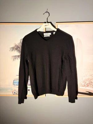 Svart v-ringad tröja Calvin Klein - Svart v-ringad tröja från Calvin Klein i mjuk ullmix. Klassisk och stilren design med ribbade muddar vid ärmslut och nederkant. Perfekt för dig som gillar enkel och clean look. Långärmad modell som passar till jeans eller chinos.