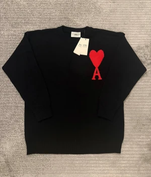 Svart stickad tröja från Ami Paris - Svart stickad tröja från Ami Paris med rött hjärta och bokstaven A broderat på bröstet. Rund hals och ribbade muddar vid ärmslut och nederkant. Tröjan har en clean och stilren look som sticker ut med den ikoniska Ami-loggan.