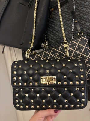 Svart axelväska med nitar - Snygg svart axelväska i quiltat skinn med guldiga nitar och kedjerem. Väskan är rymlig och är en perfekt storlek, den är aldrig använd och bra kvalitet.